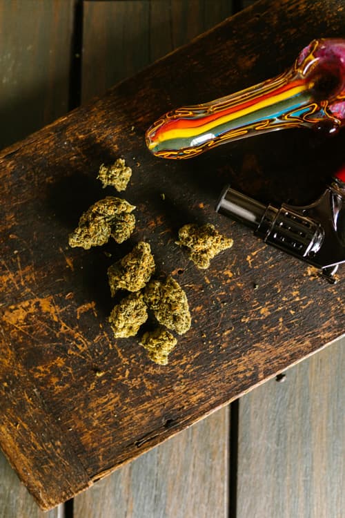 Cannabis Buds with Pipe and Lighter Ready for Use Cannabis flower, a colorful glass smoking pipe, and a torch lighter arranged on a wooden table.